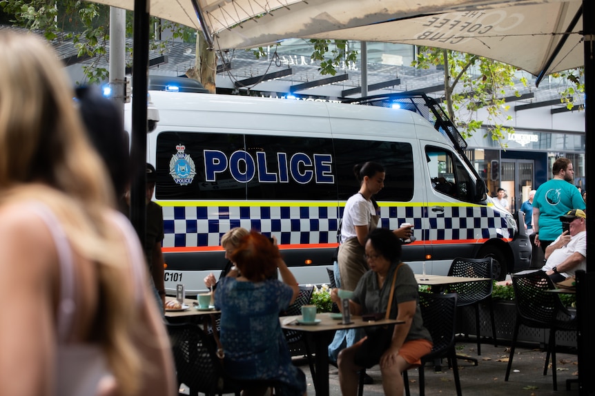 A police van drives down a pedestrian mall, past people sitting outside a coffee shop.