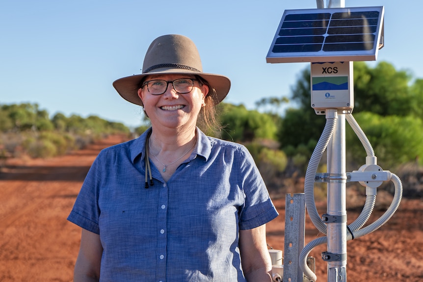 A smiling woman in a hat looks at the camera next to a small solar panel.