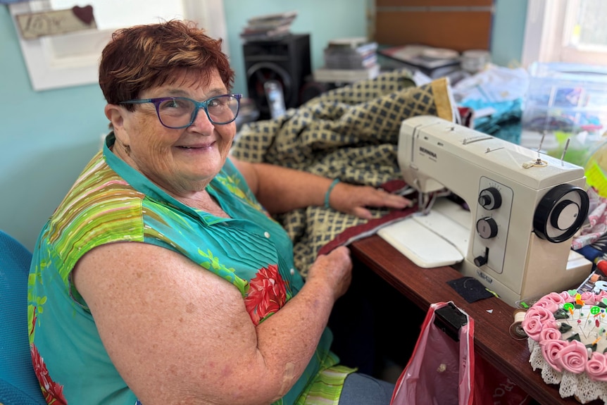 An older woman sits at a sewing machine surrounded by materials.