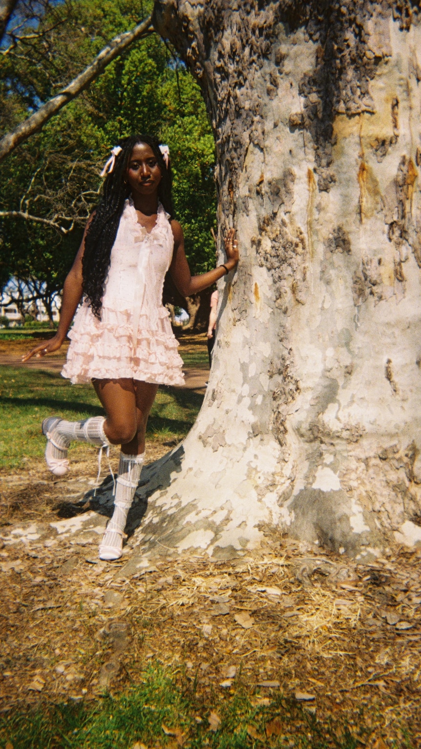 A girl with long dark pigtails wears a frilly pink dress and knee high white socks with one hand on a large tree.