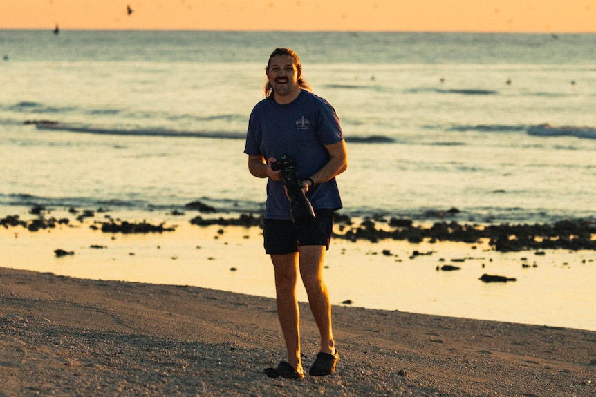 A man in his late 20s smiles standing on a beach, the sun setting behind him.