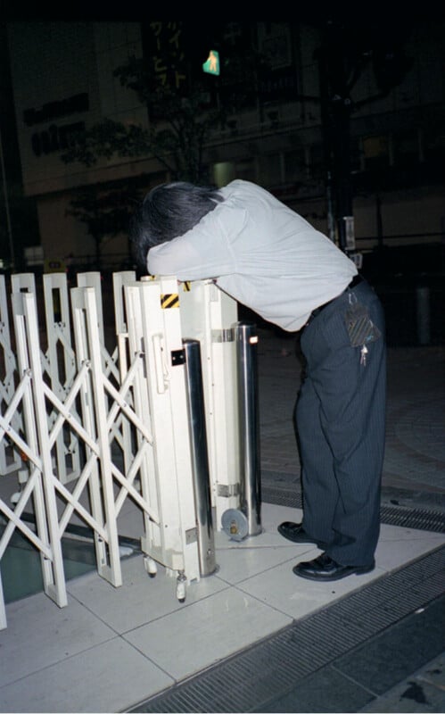 A person in business attire leans forward with their head resting on a folded metal gate at night on a city sidewalk, appearing tired or exhausted.