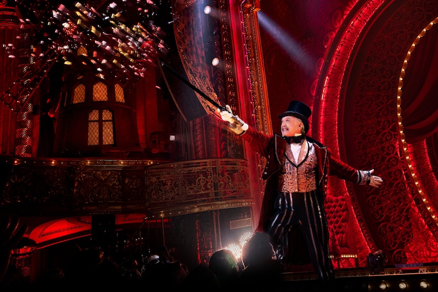 On stage, Simon Burke, early 60s, in a top hat, coat and tails, holds a cane in the air, setting off confetti.