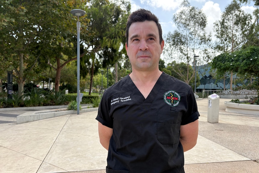 A man has short dark hair, he wears black scrubs and looks past the camera