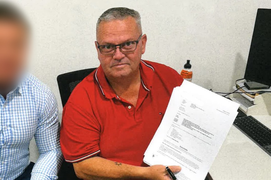 A man wearing a red polo shirt sits at a desk holding paperwork next to a person wearing a blue checkered shirt.