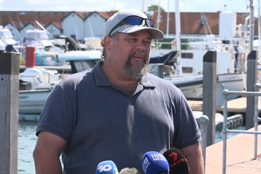 A man wearing a cap speaks in front of microphones at a boat harbour.