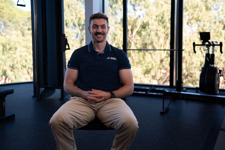 A man in a navy polo shirt and beige trousers smiles. Gym equipment is in the background.