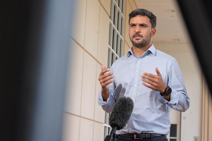 A man with facial hair, wearing a serious facial expression and gesturing with his hands as he speaks into a news microphone.