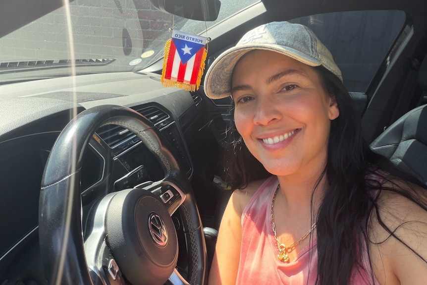 Woman sitting in a car with sunlight streaming in, a Puerto Rican flag hanging from the mirror.