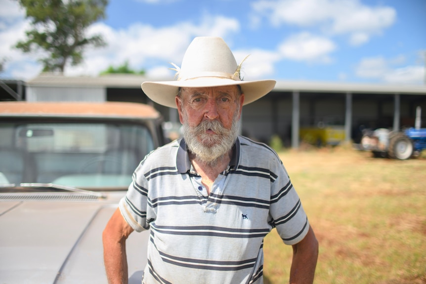 An older man whith a white beard and a cowboy hat stands in-front of a large open sided shed. 