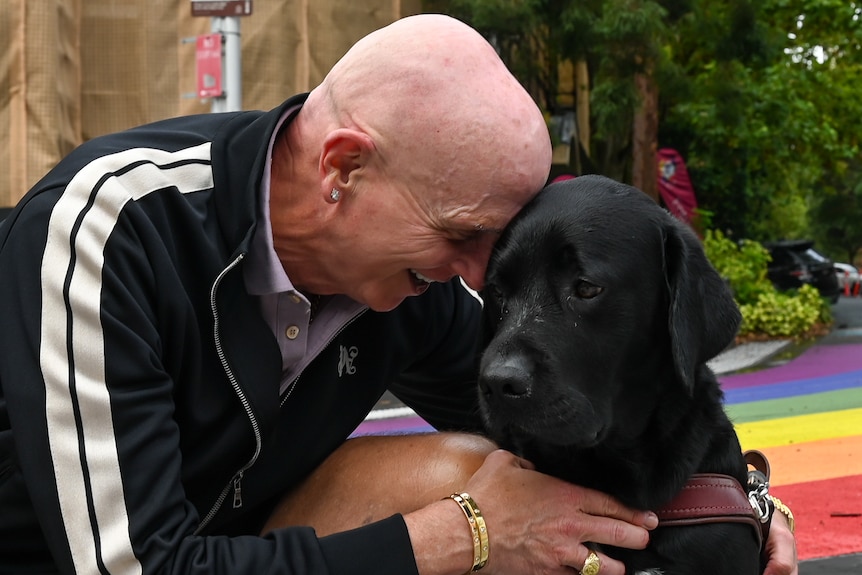 A man in a black tracksuit jumper hugs a black labrador guide dog.