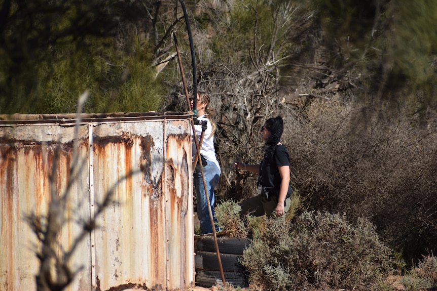 A detective on a ladder looking into a large rusty water tank as another detective looks on in scrubland