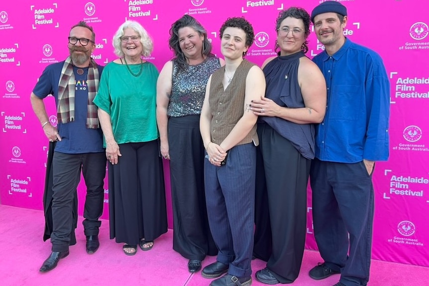 Six people pose and smile standing on a pink carpet against a pink media wall that reads 'Adelaide Film Festival'