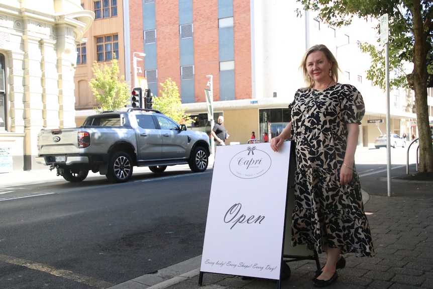 A woman stands next to an A-frame sign in city street.