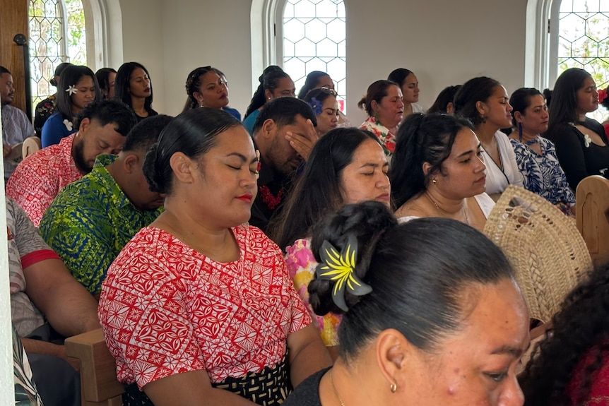 Men and women sit closely together in wooden pews inside a small chapel.