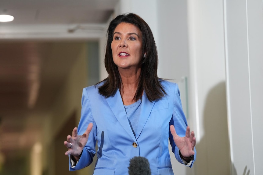 A woman wearing a pale blue suit jacket and blouse, speaking in a corridor.