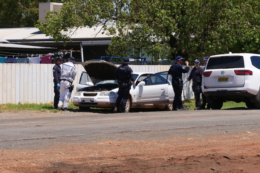 A group of police officers surround an abandoned car parked near a tin fence.