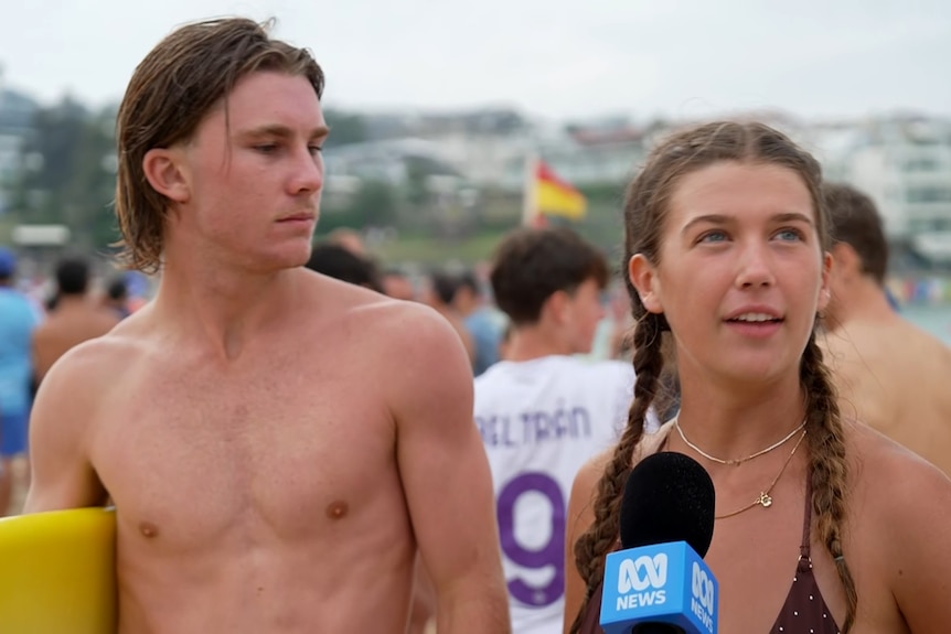 A man holding a yellow surfboard stands next to a woman with her hair in braids speaking into an ABC microphone.