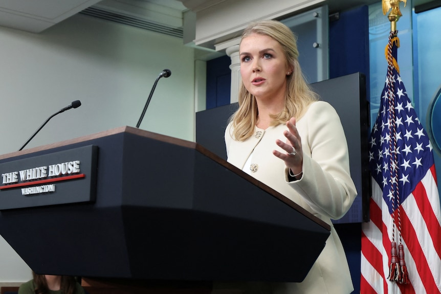 Karoline Leavitt standing at a podium with two microphones facing her.