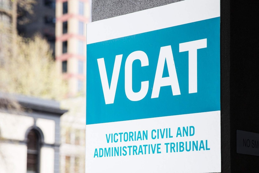 A blue and white sign is attached to a concrete pillar with a streetscape in the background.