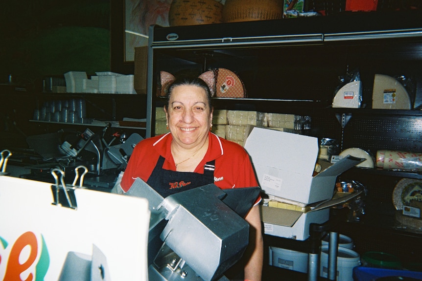 A woman in an apron smiles as she stands in front of a deli slicer, in front of shelves of cheese.