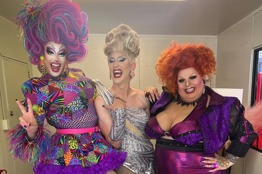 Three drag queens dressed in purple and silver pose.