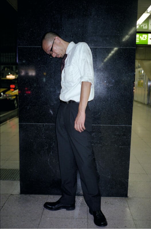 A man in glasses, a white shirt, and dark trousers stands indoors, leaning with his head down and arms hanging loosely, against a shiny black wall.