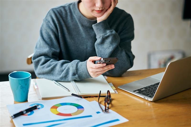 Young woman rests her head in her hands on a desk while looking at her phone. 
