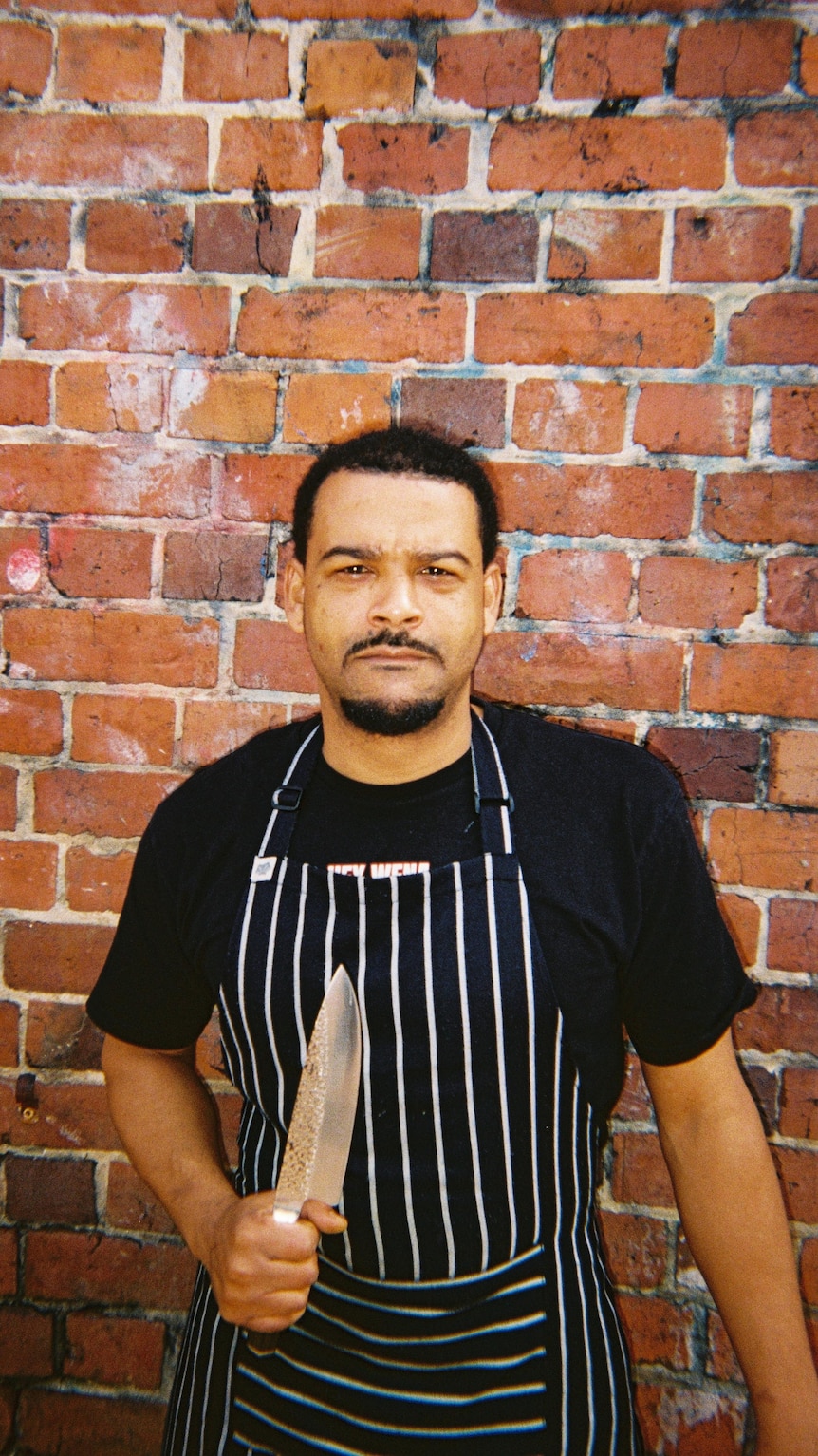 A man in a striped apron stands in front of a brick wall and holds a chef's knife up.