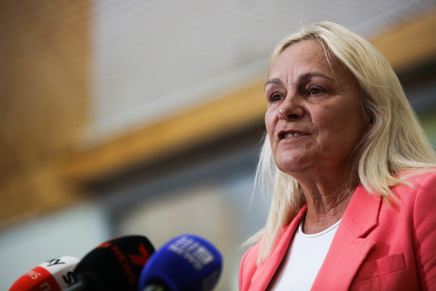 A close-up side-on shot of Sabine Winton speaking into microphones at a media conference, wearing a pink jacket and white top.