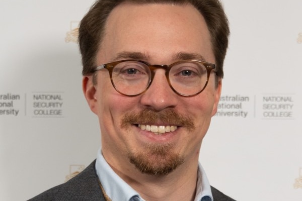 A man with brown hair and facial hair wearing glasses smiles
