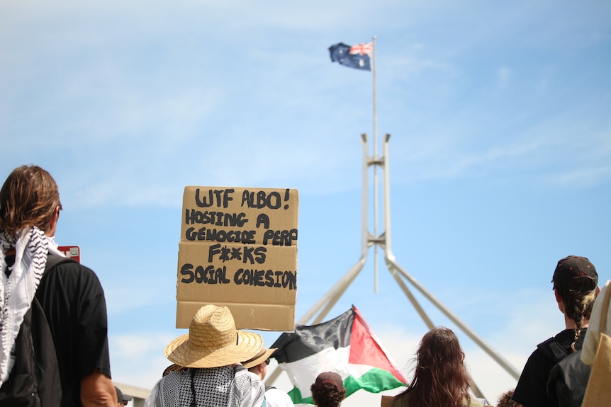 Protesters holding placards, as the Australian flag flies in the background over Parliament House.