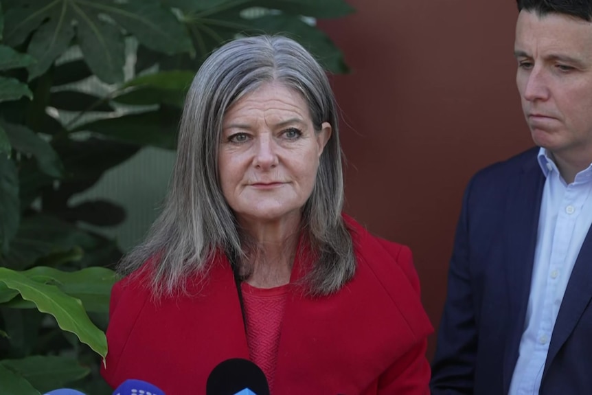 A woman wearing a bright red jacket stands in front of media microphones.