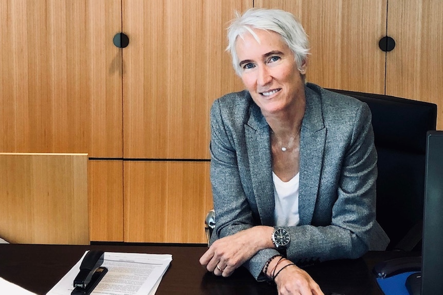 A formally-dressed woman with grey short hair sits in a wood-panelled office.
