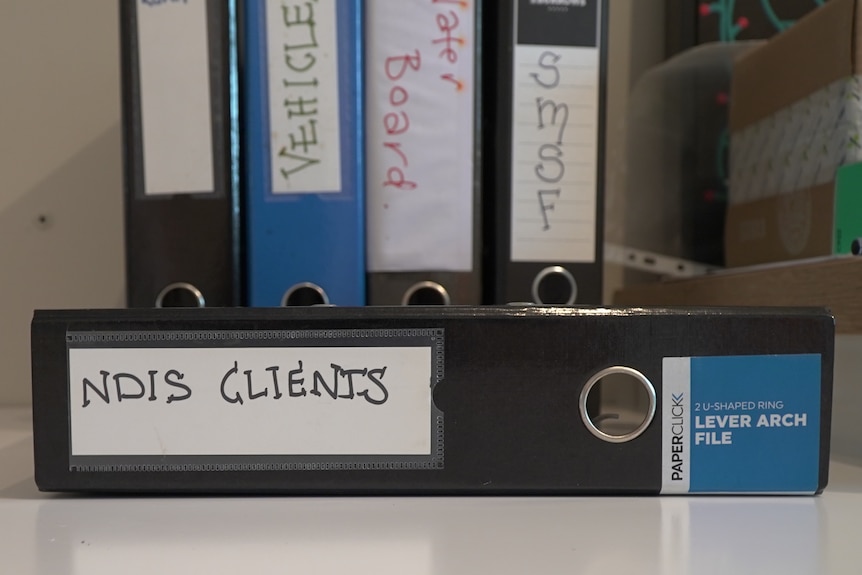 A stack of folders on a shelf with one folder reading 'NDIS clients'.