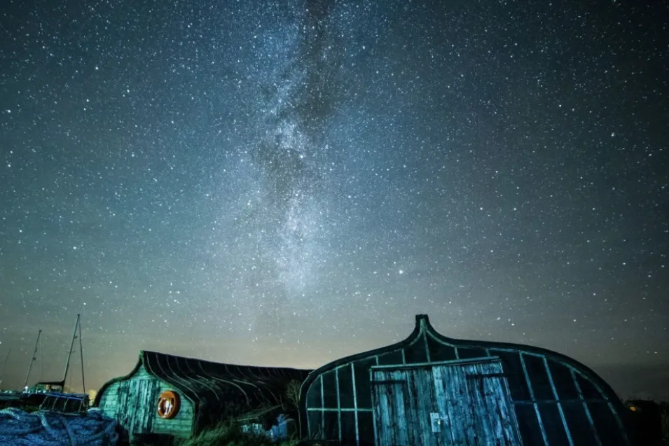 The sky at night is dotted with bright stars and a streak of black that looks like a tear in the sky. There are single-storey wooden huts in the foreground. The roofs are curved up to a single point and there are small ships moored on the left.