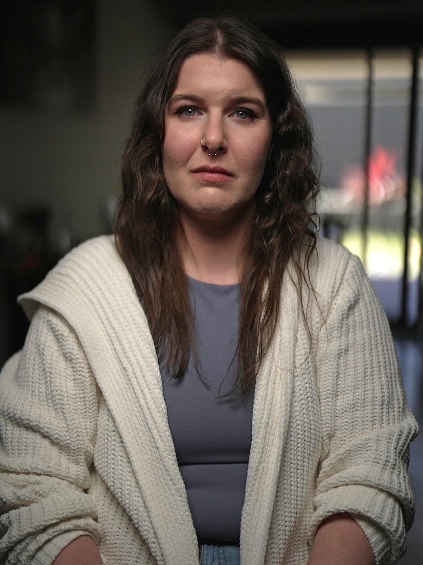 A woman sits indoors, looking at the camera with a sad, serious expression.