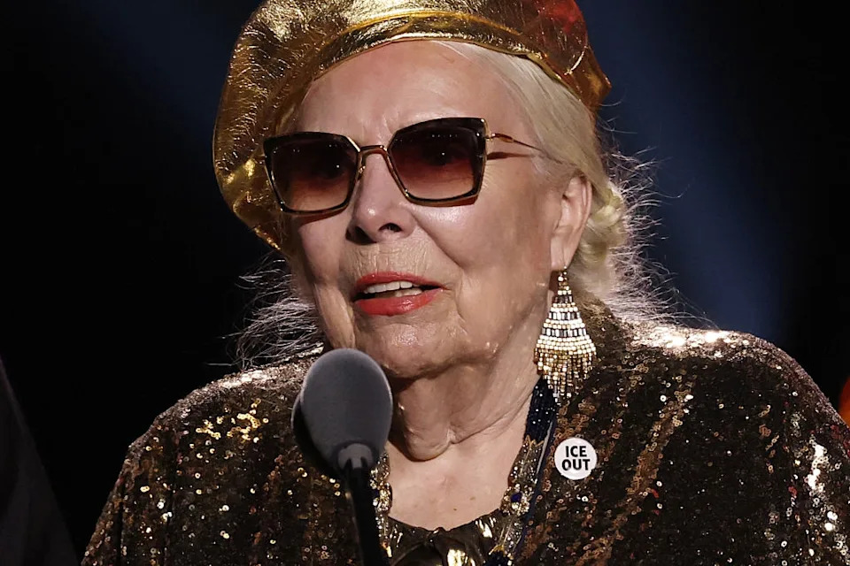 A person on stage wearing sunglasses, a sequin outfit, and a gold beret, with a statement pin reading "Ice Out."