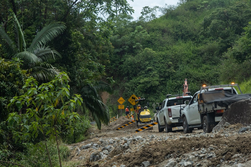 Utes line up behind a small digger, road barriers in place across the road in the middle of a rainforest