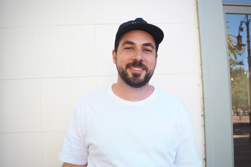 A young man with a beard smiles at the camera. He is wearing a cap and t-shirt. 