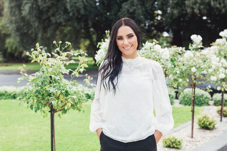 a woman in a white shirt smiling in front of plants and flowers