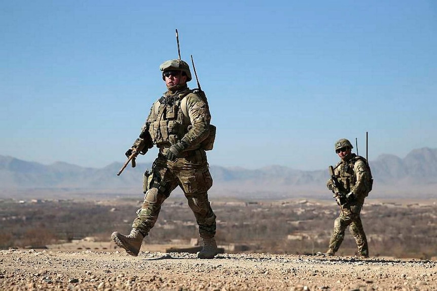 Soldiers with weapons walk through a barren landscape.