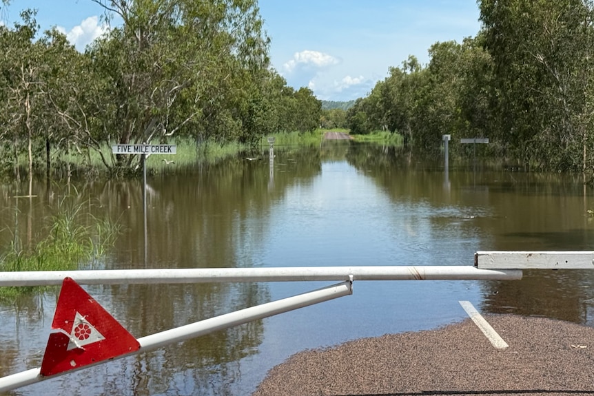 A road with white pole blocking it, water flooding a majority of the road, sign under water reads 'five mile creek'