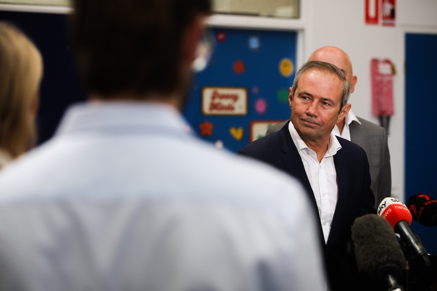 A mid-shot of WA Premier Roger Cook speaking into microphones at a media conference, with a man back turned in the foreground.