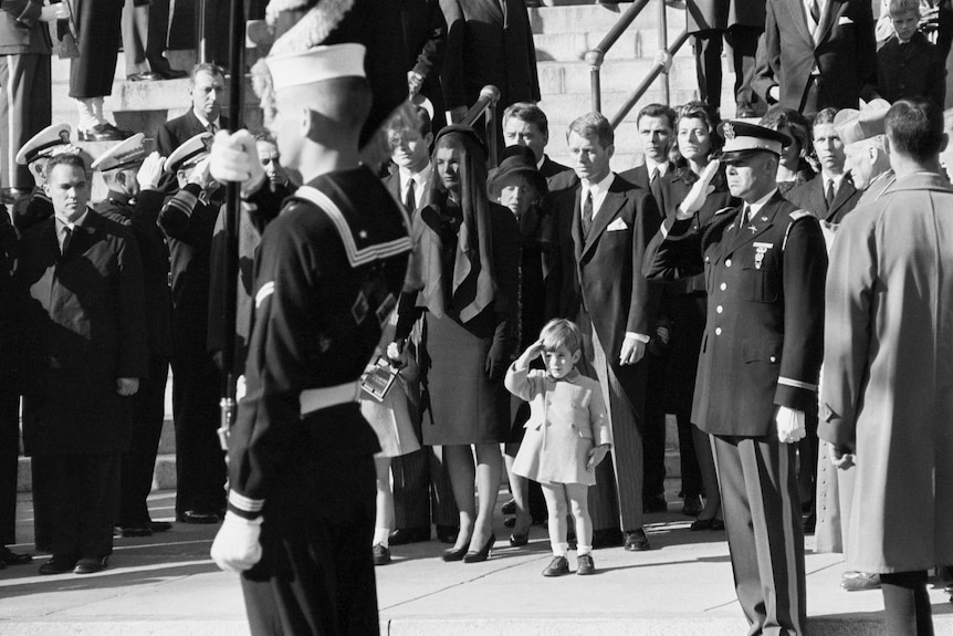 A black-and-white photo of a little boy saluting with his right hand as a funeral procession passes.