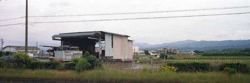 A large white industrial building stands amid overgrown grass and bushes, with power lines and mountains visible in the background under a cloudy sky.