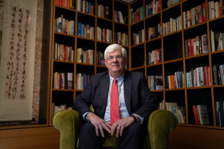 A man with glasses and a suit and tie sits in a light green chair surrounded by books in his personal library.