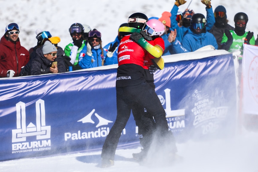 A pair of snowboarders embrace on a snow track in front of a crowd.