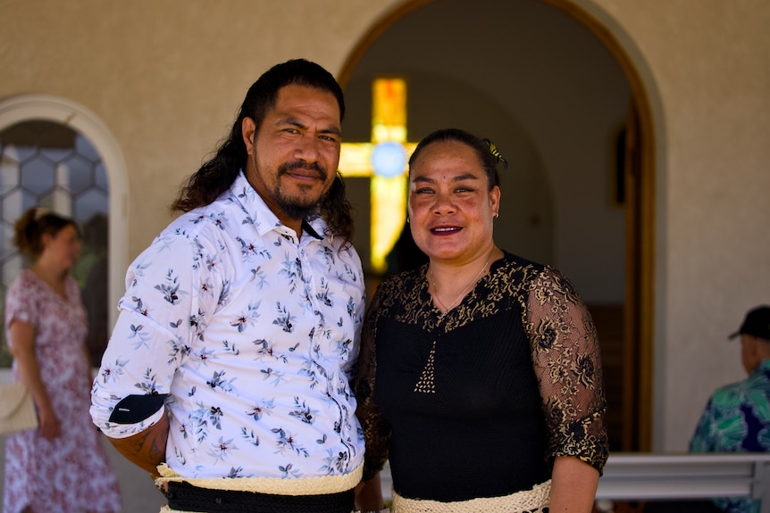 A man and woman stand outside a small chapel with a stained-glass cross glowing behind them.