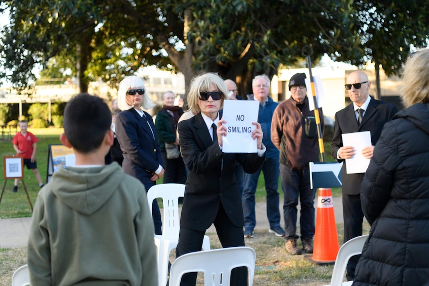 People dressed in black suits hold a sign telling people there is no smiling